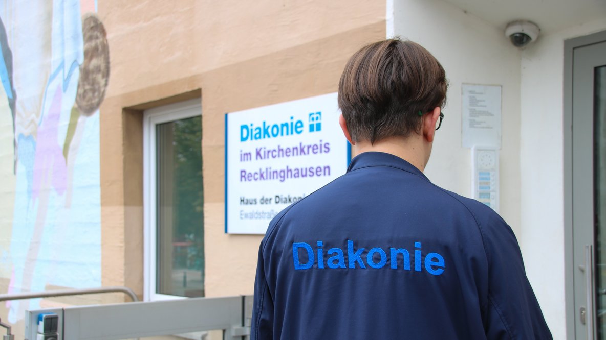 Ein Pfleger steht mit dem Rücken zur Kamera, auf dem Rücken steht Diakonie