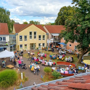 Außenansicht von oben auf Haus Jona und den parkähnlichen Garten. Man sieht ein Sommerfest. es ist geschmückt und viele Menschen sind draußen.