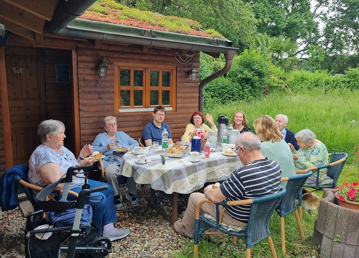 Kaffeerunde vor einem Holzhaus mit Grasdach, im Hintergrund ist eine hochgewachsene Wiese