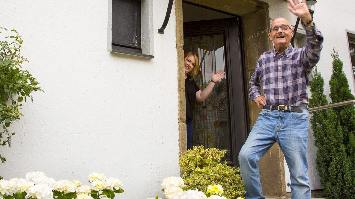 Mann steht an der Eingangstür des Hauses und winkt. auch eine Frau winkt aus der Eingangstür heraus 