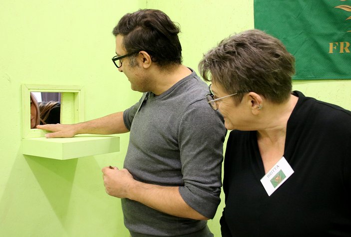 Demiz Tekmen und Britta Porbatnik-Schmelter schauen gebannt auf eine Klappe in der Wand, durch die eine Hand Befehle an die Asylsuchenden gibt