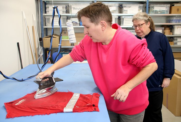 Eine Frau im pinkfarbenen Pulli bügelt ein rotes Stück Stoff, beobachtet von einer Kollegin.