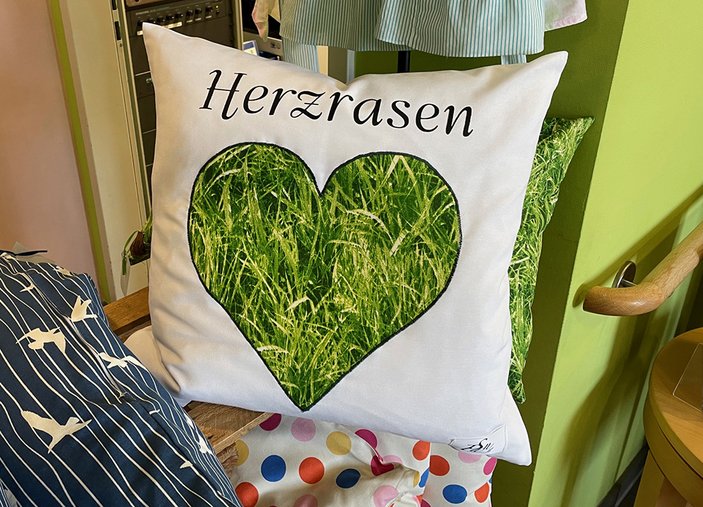 Kissen mit einem grünen Herz, das Rasen in Nahaufnahme zeigt. Dazu die Überschrift: Herzrasen