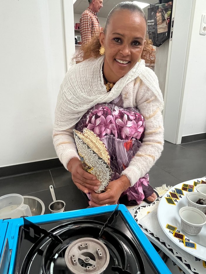 Eine am Boden knieende Frau in einem rosa geblümten Kleid füllt Kaffeemehl mit einem geflochtenen Trichter in einen Tonkrug