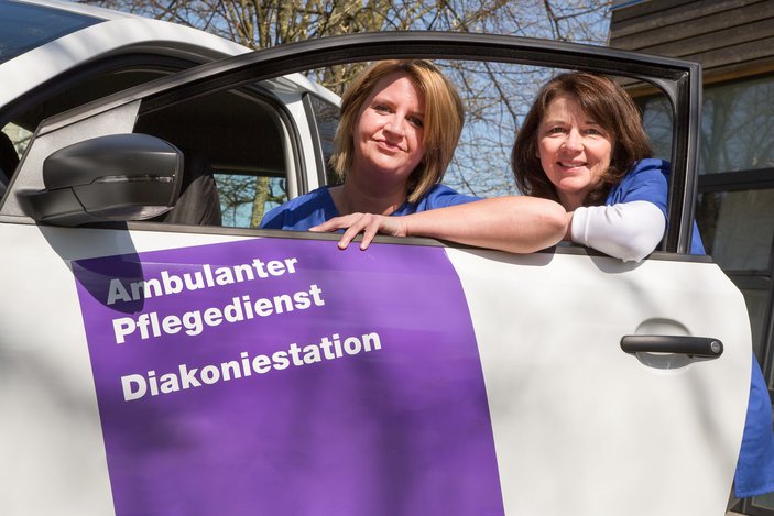 Zwei Frauen lehnen sich an eine Autotür eines Pflegedienstes und lächeln