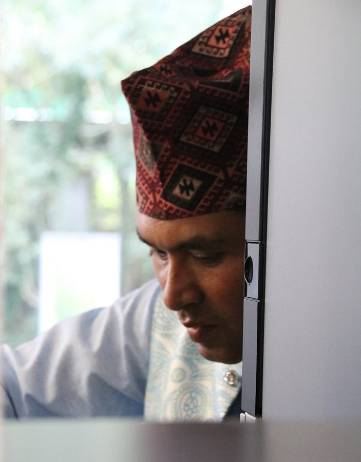 Portrait des Koches mit einer traditionell nepalesischen Mütze, durch ein metallenes Küchenregal hindurch fotografiertchenregal 