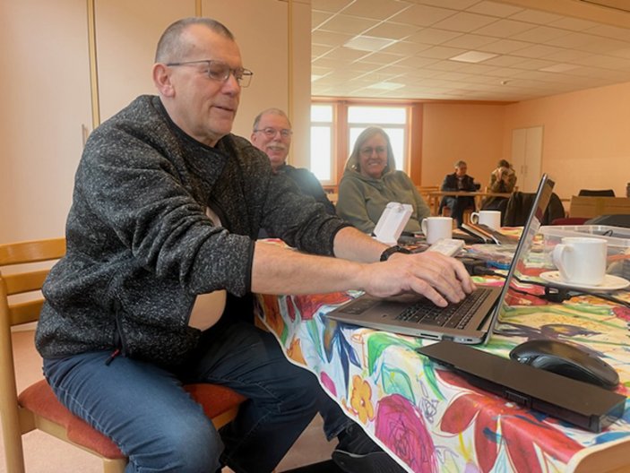 Mann an einem Tisch mit bunter Decke, der an einem Laptop arbeitet, beobachtet von einem Paar im Hintergrund