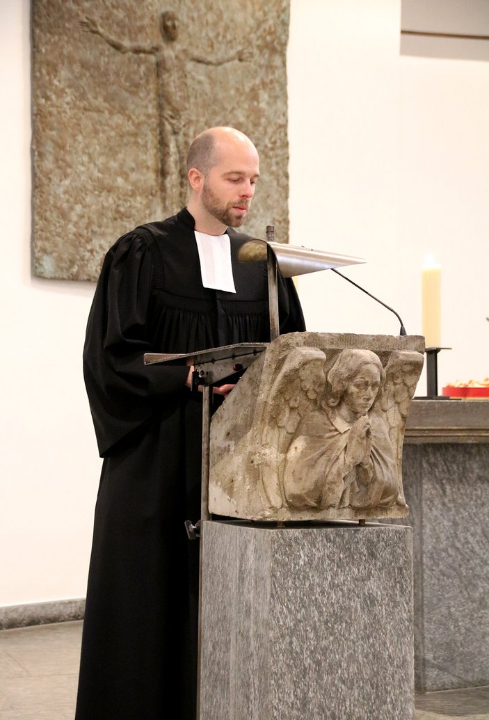 Der Pfarrer im Talar am Rednerpult, das von einem steinernen Engel verziert ist. Im Hintergrund eine moderte Christusdarstellung in einem Gemälde 
