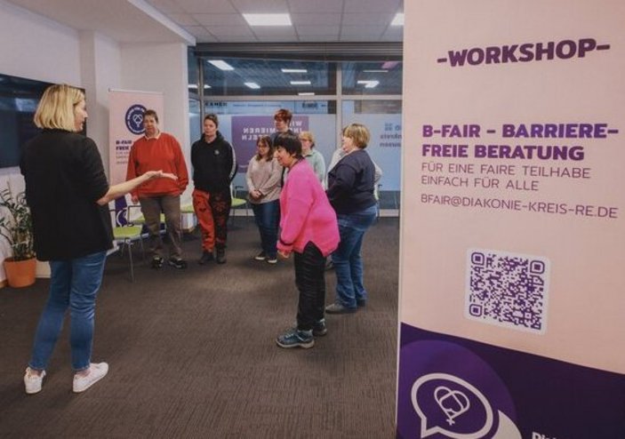 Man sieht Personen bei einem Workshop in einem Raum, Im Vordergrund steht ein Roll-Up