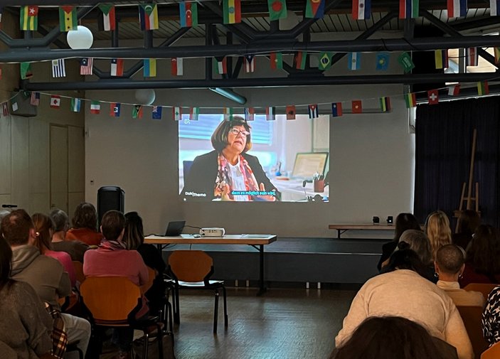 Große Leinwand im dunklen Raum, auf der ein Film läuft. Zuschauer*innen von hinten