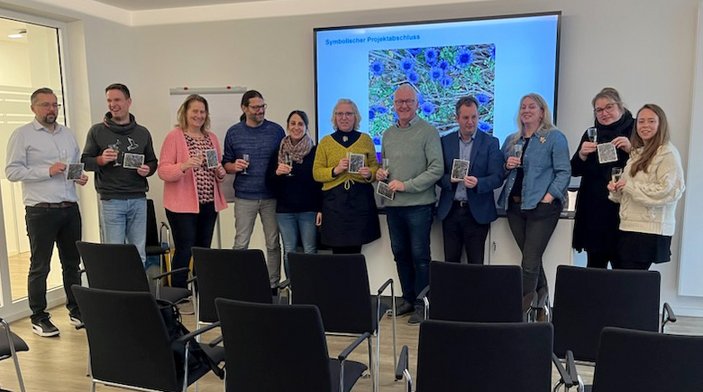 Gruppenfoto vor Bildschirm, die Abgebildeten halten Sektgläser in ihren Händen