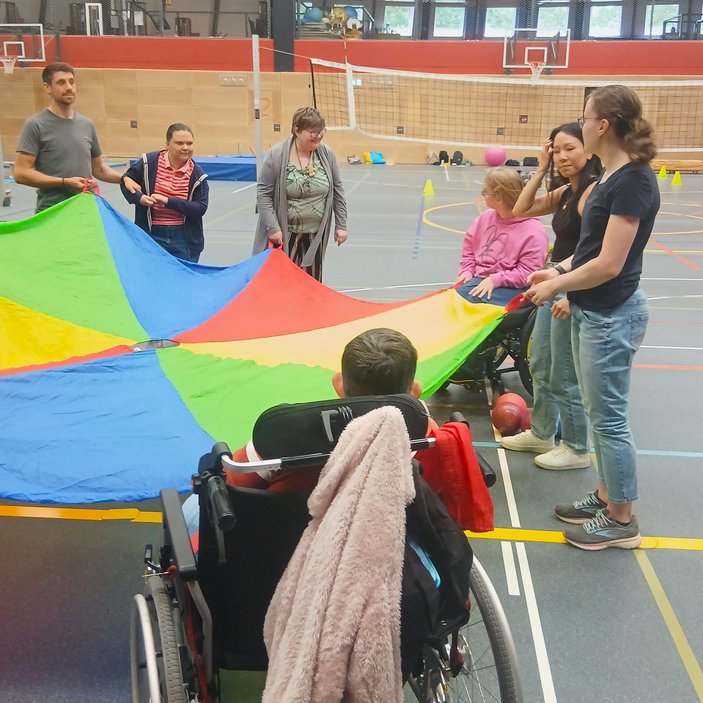 Mehrere Personen halten ein buntes Tuch in einer Turnhalle. manche von ihnen sitzen im Rollstuhl, manche stehen