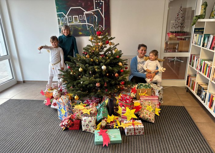 geschmückter Weihnachtsbaum im Stadtteiltreffen, darunter viele bunt verpackte Geschenke, die Einrichtungsleiterin Ramona Glodschei, eine Mutter und ihre zwei als Engel verkleideten Kinder